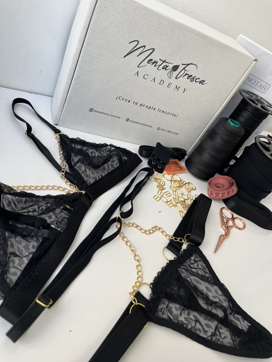 Conjunto de encaje negro con cadenas doradas, junto a la caja del taller, hilos y herrajes.
